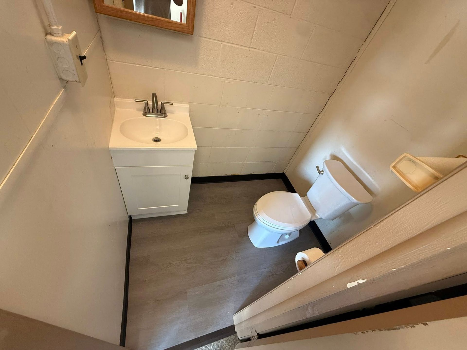 A small, high-angle view of a bathroom with a white pedestal sink, a white toilet, and gray wood-look flooring.