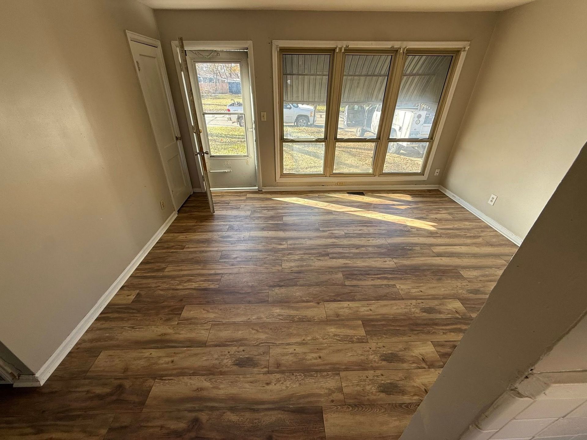An empty room featuring light-colored walls, brown wood-grain flooring, a glass-paned door, and large windows.