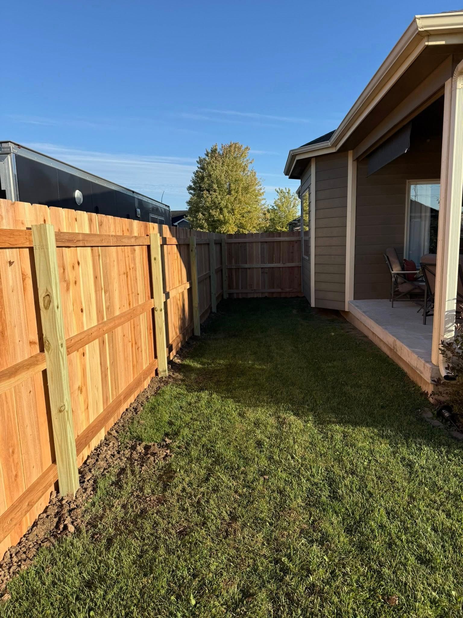 A newly installed wooden fence runs along the side of a house, bordering a lawn leading to a patio under a roof overhang.