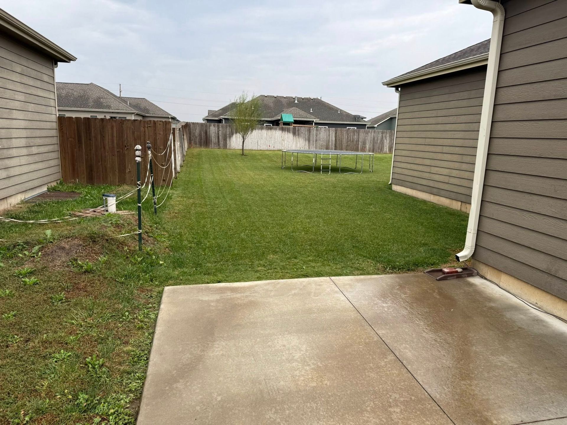 A backyard view featuring a concrete patio, a green lawn, wooden fences, and houses on both sides.