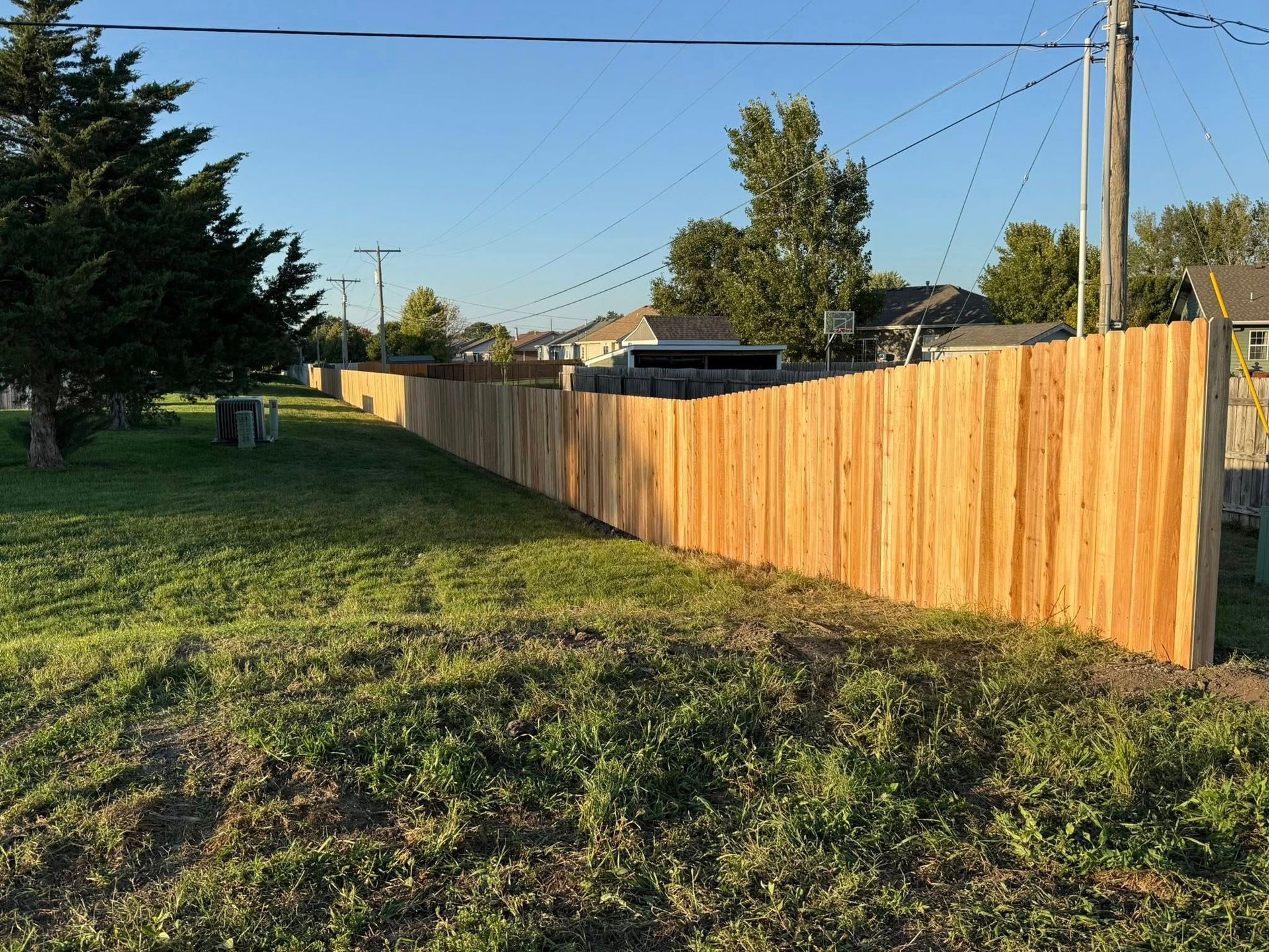 A long, newly installed wooden privacy fence runs across a grassy yard under a clear blue sky.