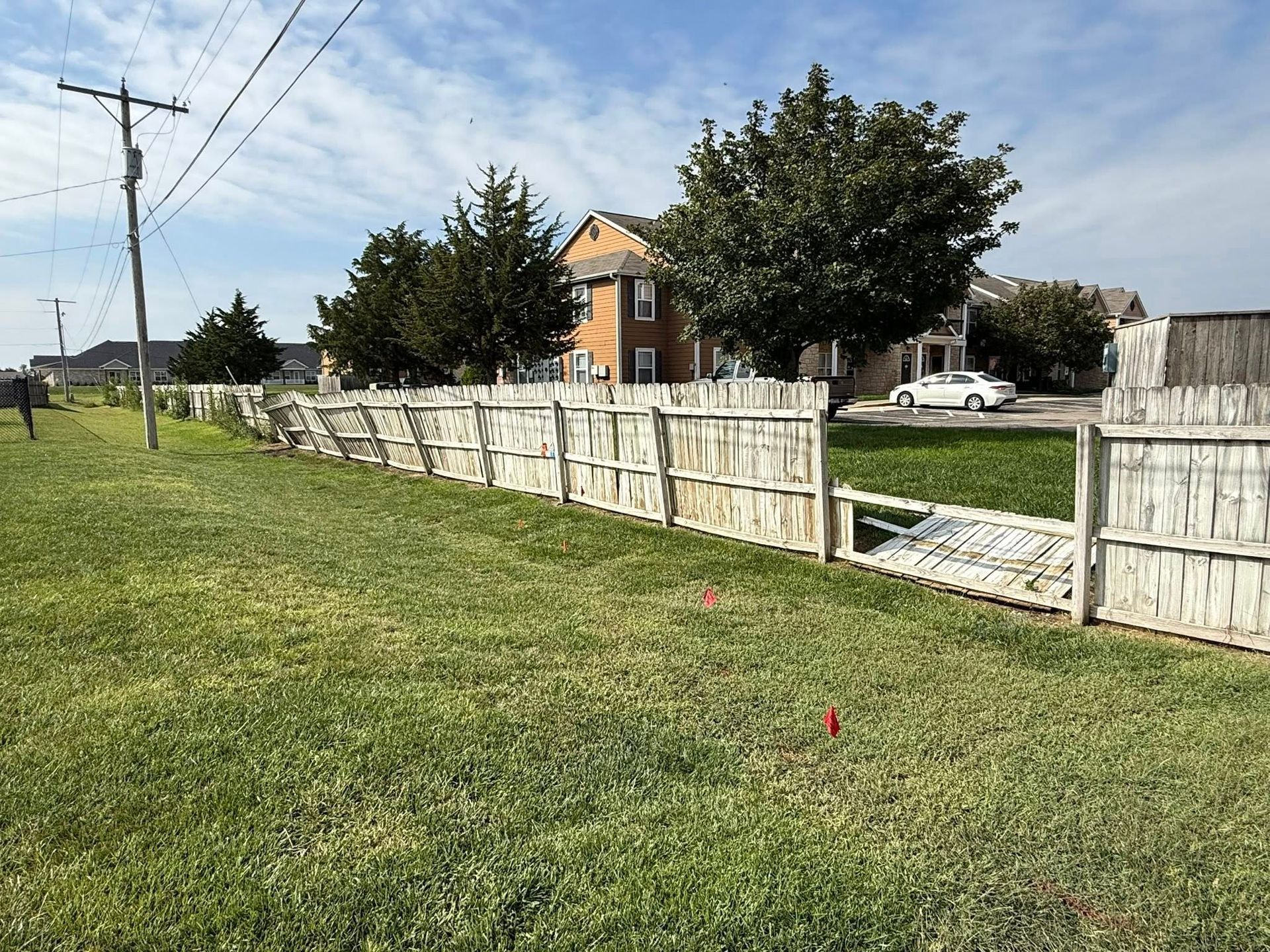 A weathered, damaged wooden fence borders a grassy yard with trees and a house in the background under a blue sky.