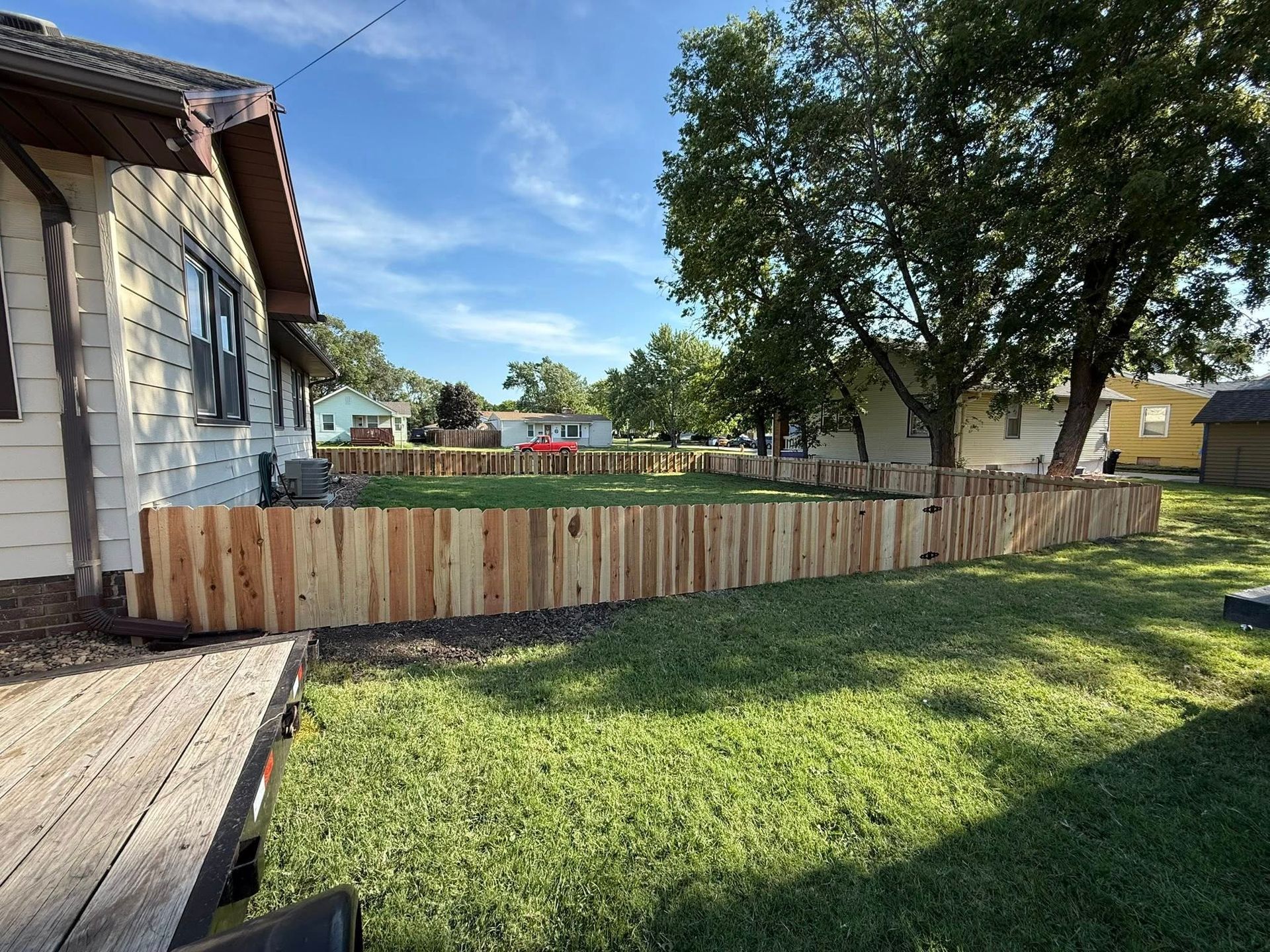 A newly installed wooden picket fence encloses a green backyard next to a cream-colored house on a sunny day.