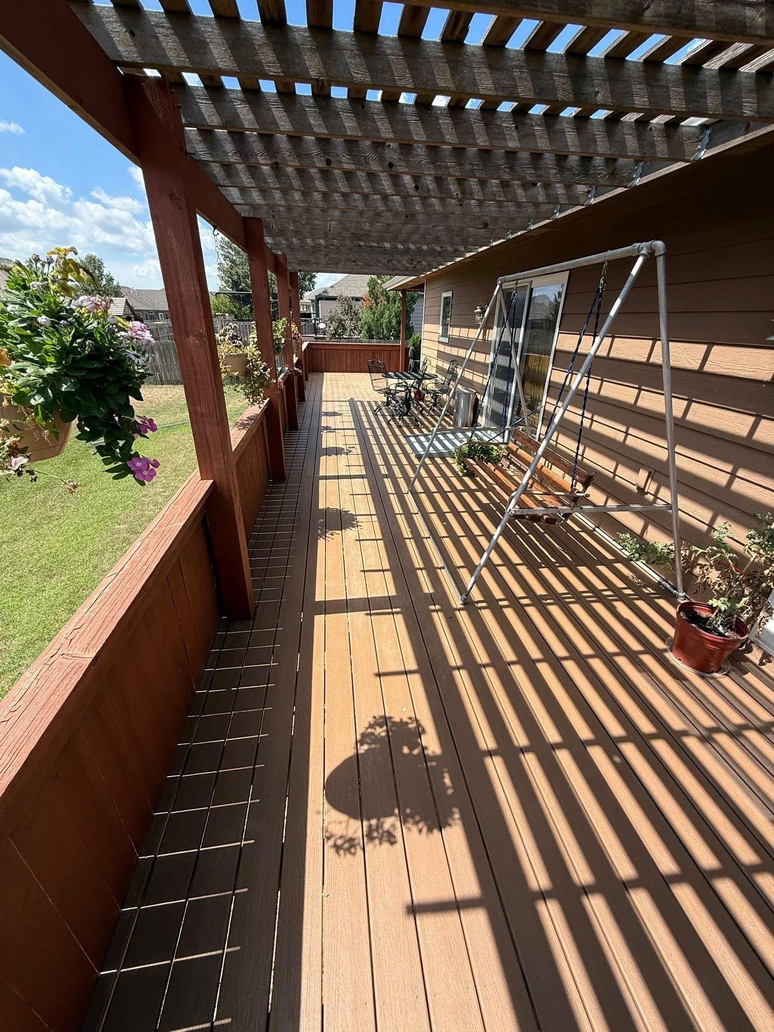 A wooden deck with a pergola roof casts shadows across the floor, featuring a swing set, potted plants, and a brick wall.