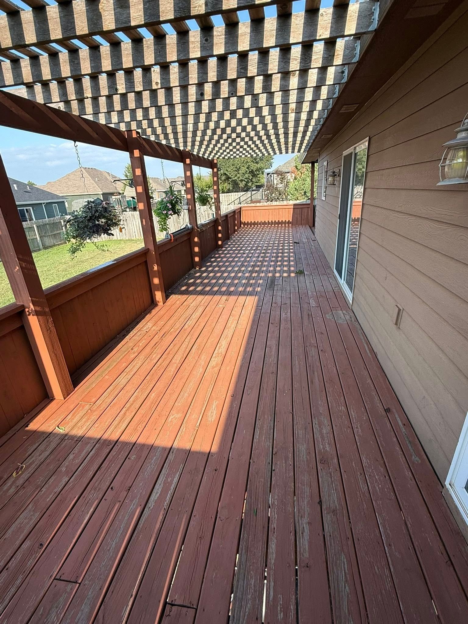 A wooden deck with a slatted pergola roof, featuring red-stained planks and a wooden railing overlooking a grassy yard.