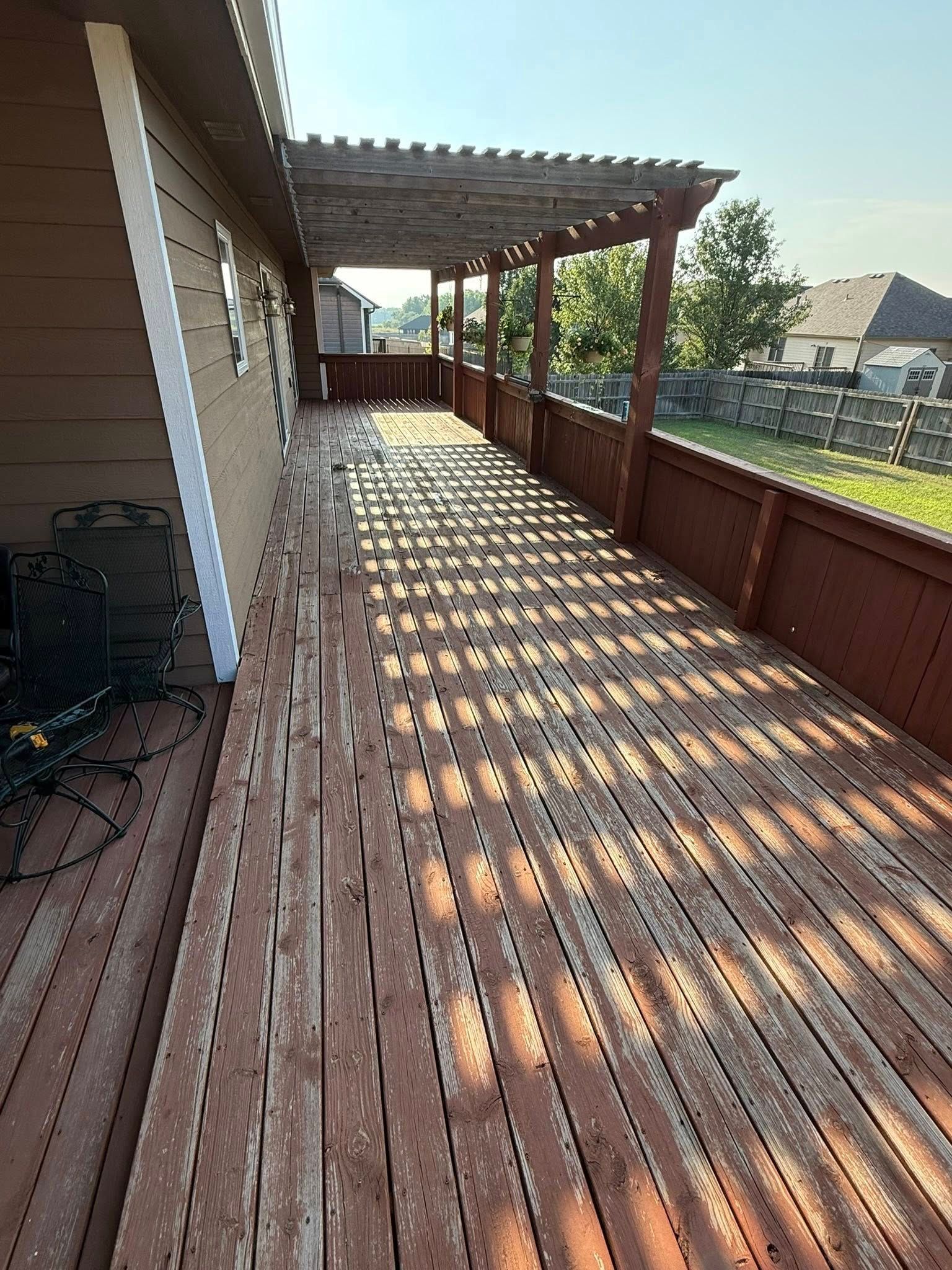 A long, weathered brown wooden deck with a slatted pergola roof stretches alongside a house wall in daylight.