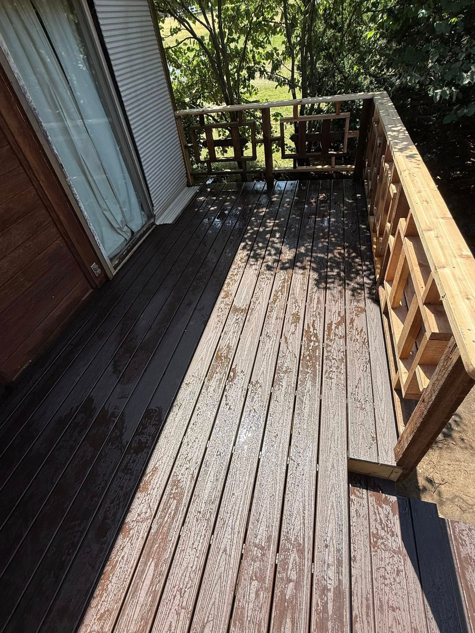 An outdoor deck with faded, weathered wooden planks leading to a railing, partially shaded by sunlight and trees.
