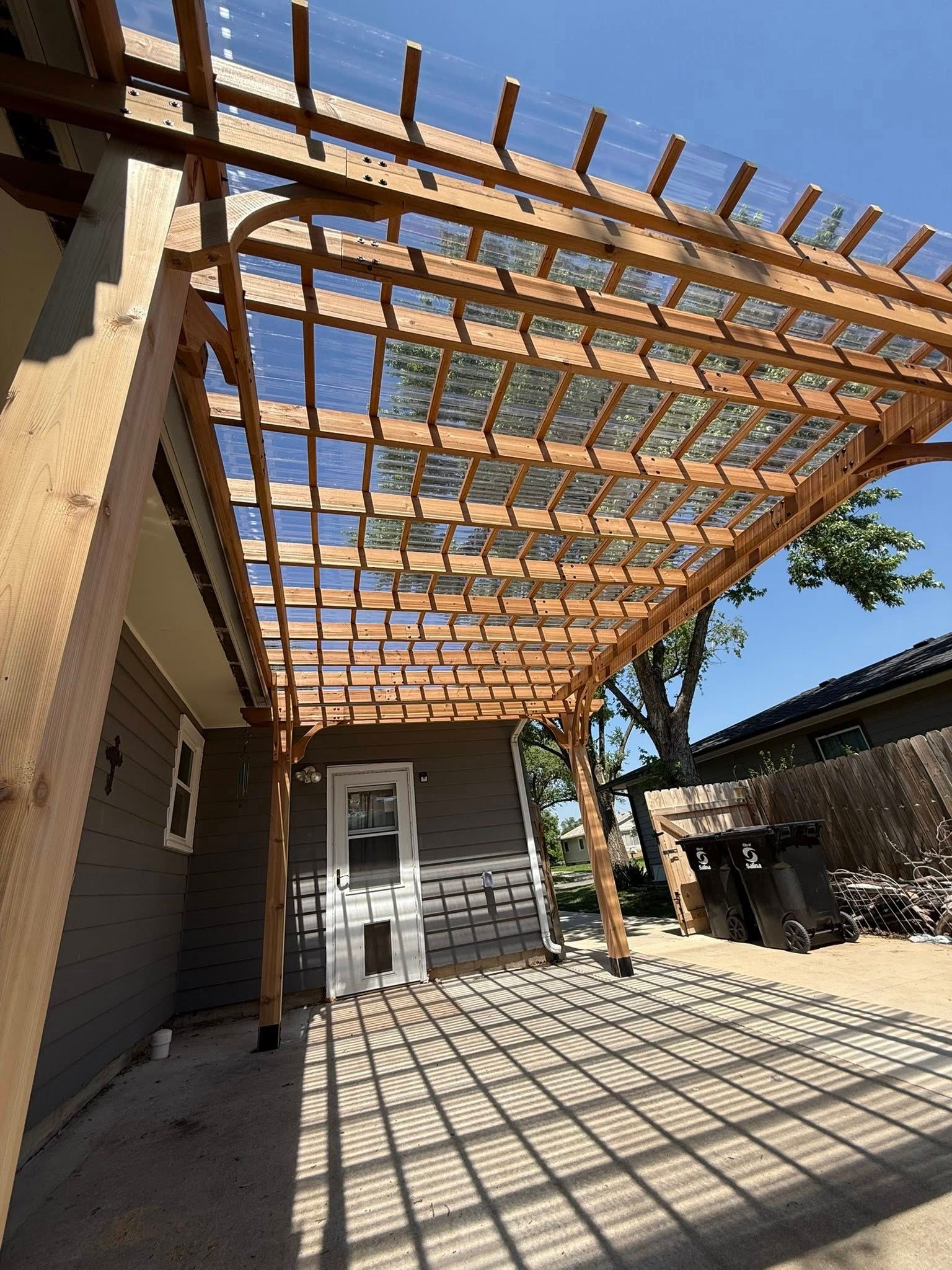 A wooden pergola with a clear, corrugated plastic roof attached to the side of a grey house with a white door.
