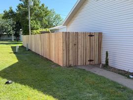 A new vertical cedar wood fence extends from the side of a white-sided house toward a green lawn and trees.