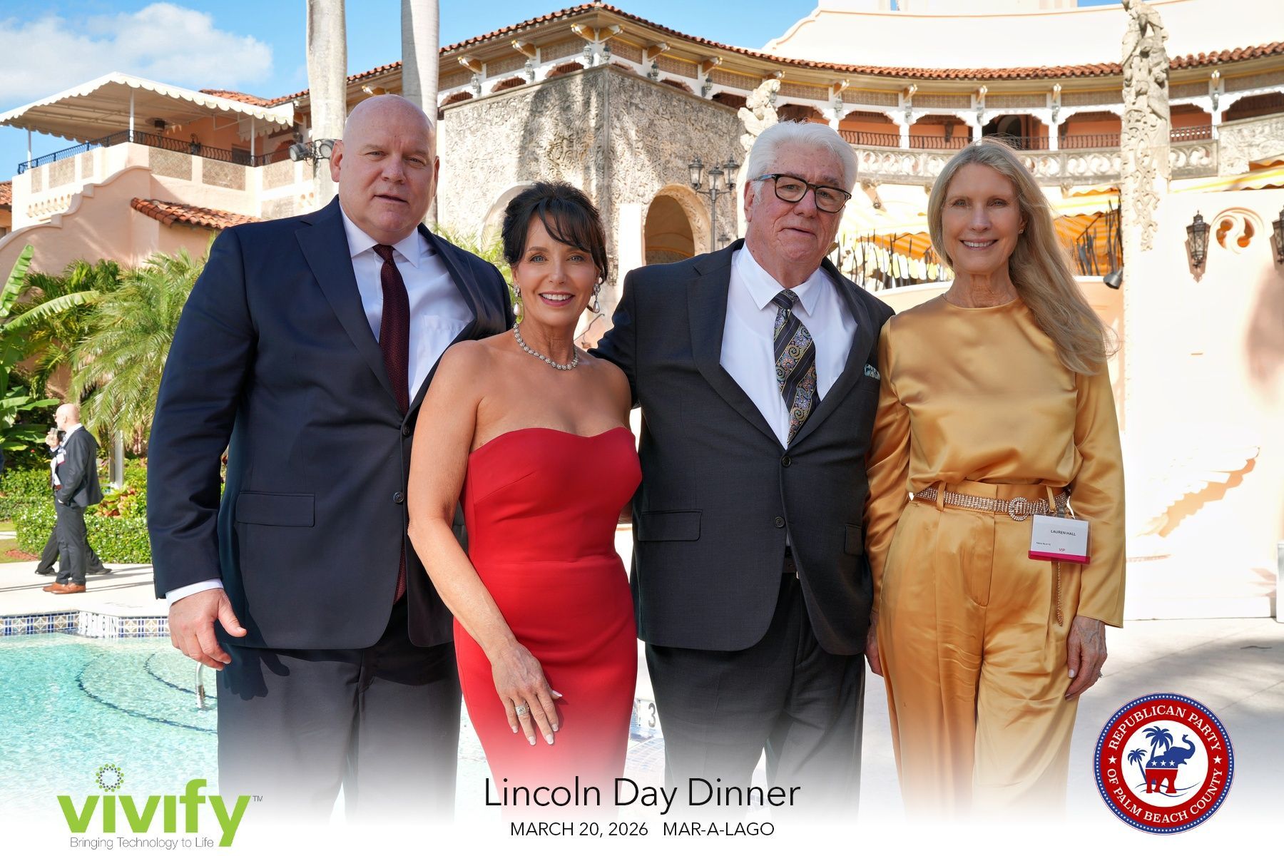 Four people posing outdoors by a pool at a formal event, with ornate buildings in the background.