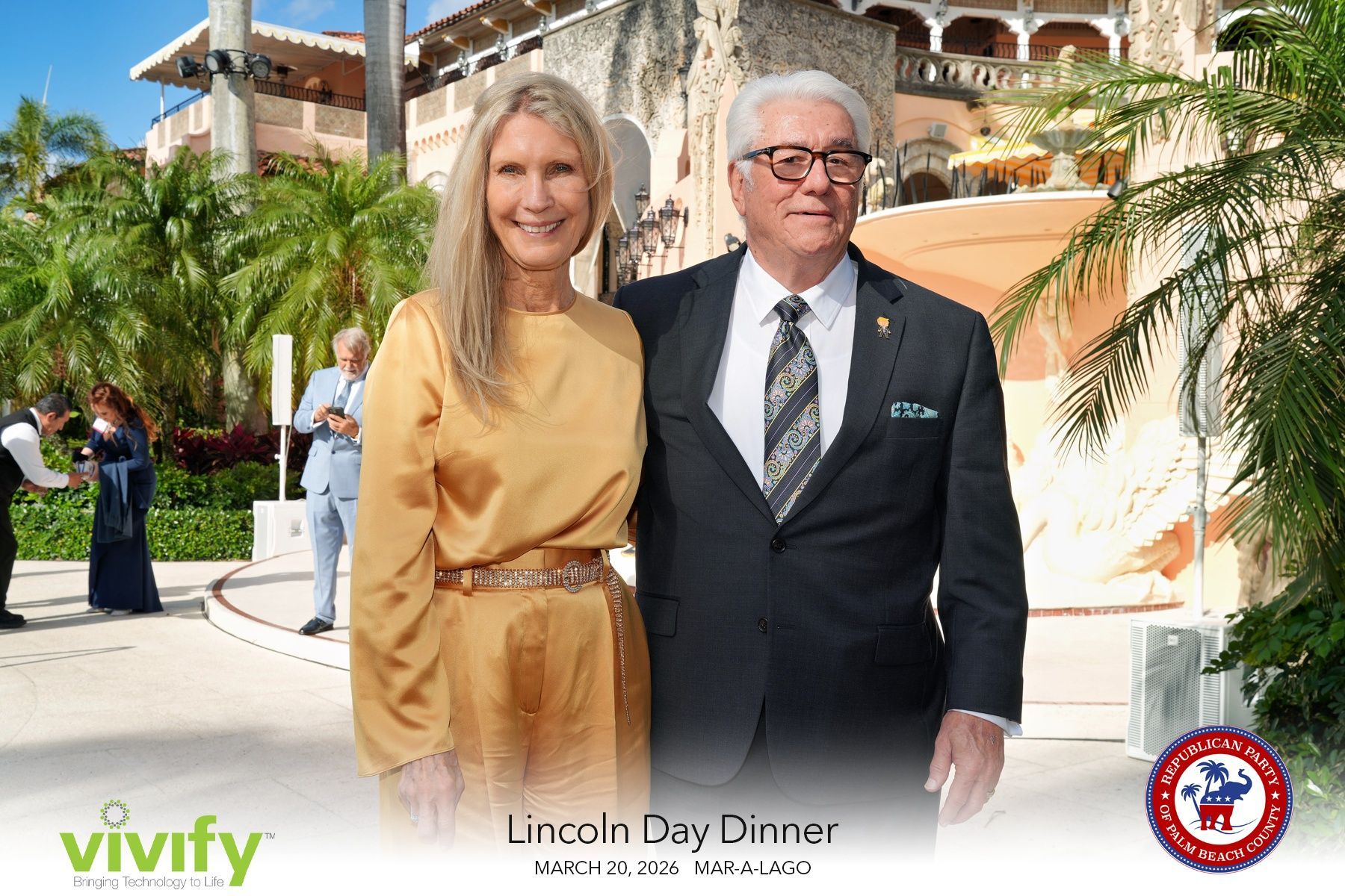 Two adults posing outdoors in formal attire at a sunny event venue, with palm trees and a building behind them.