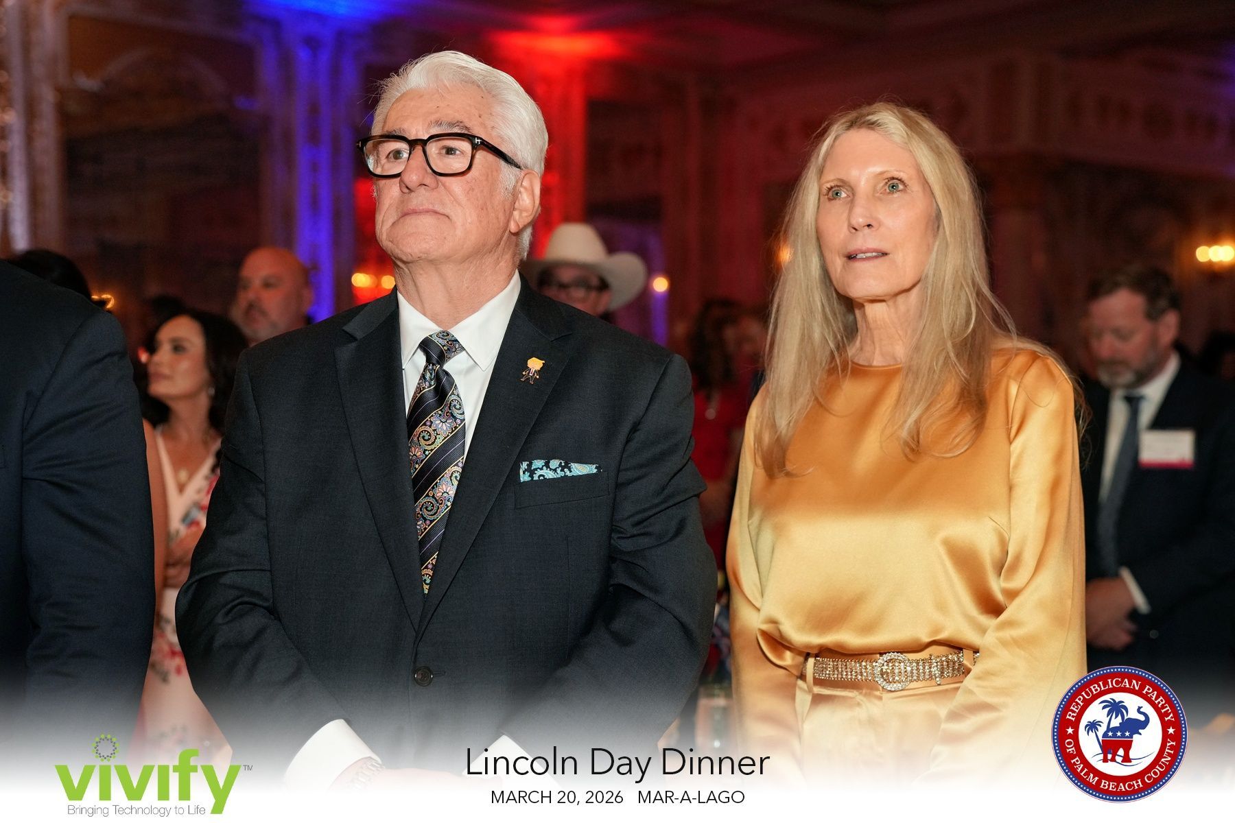 Two formally dressed guests seated at a gala dinner with red ambient lighting and event branding.