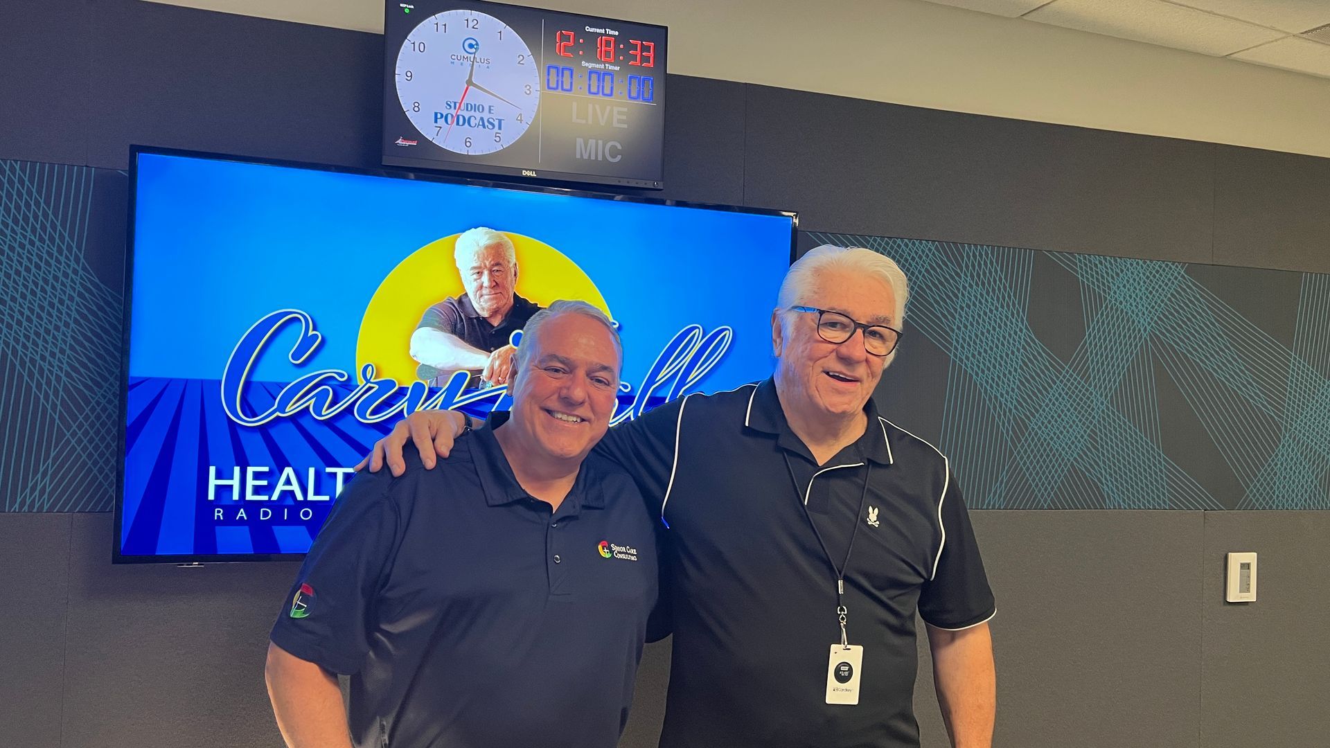 Two men in dark polos standing in a room in front of a blue screen and wall clock