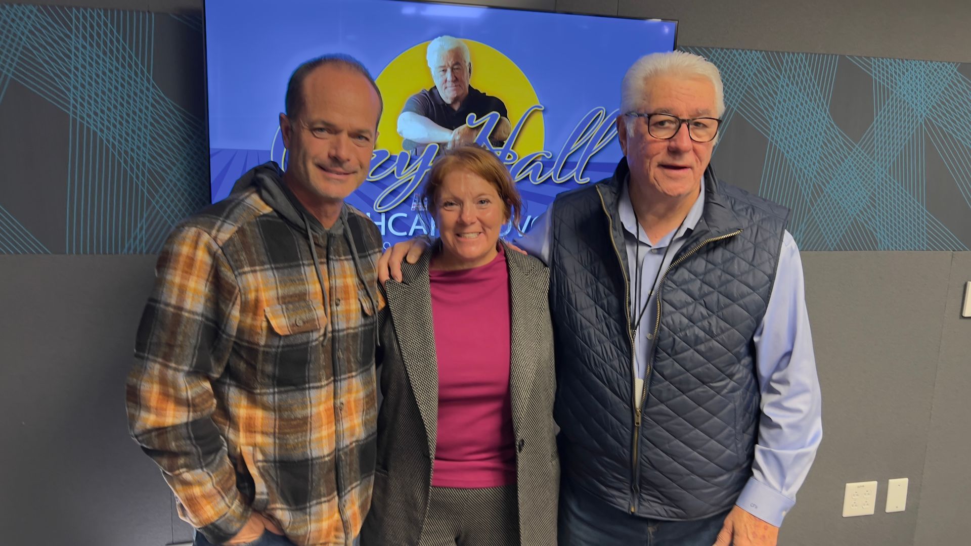 Three people smiling indoors in front of a screen, wearing plaid, pink, and a gray vest.
