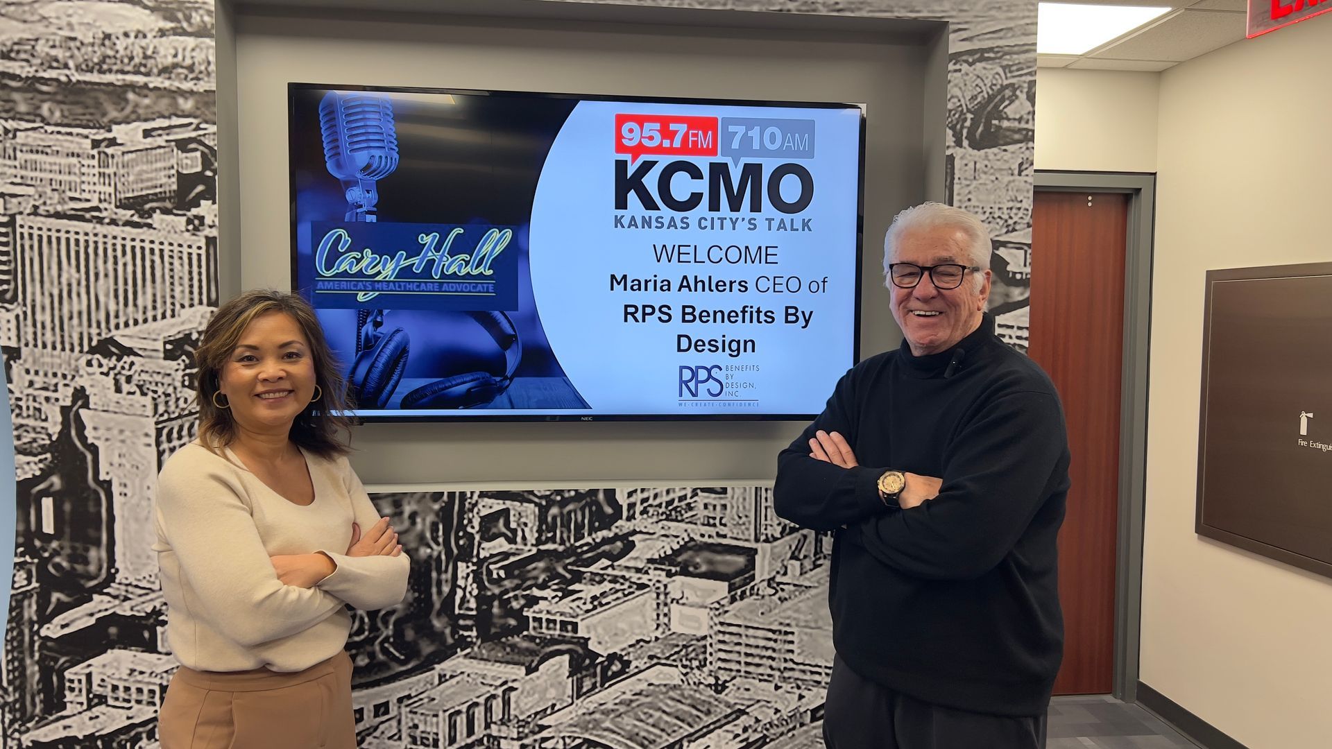 Two people posing beside a wall-mounted TV showing a KCMO radio station screen in an office.
