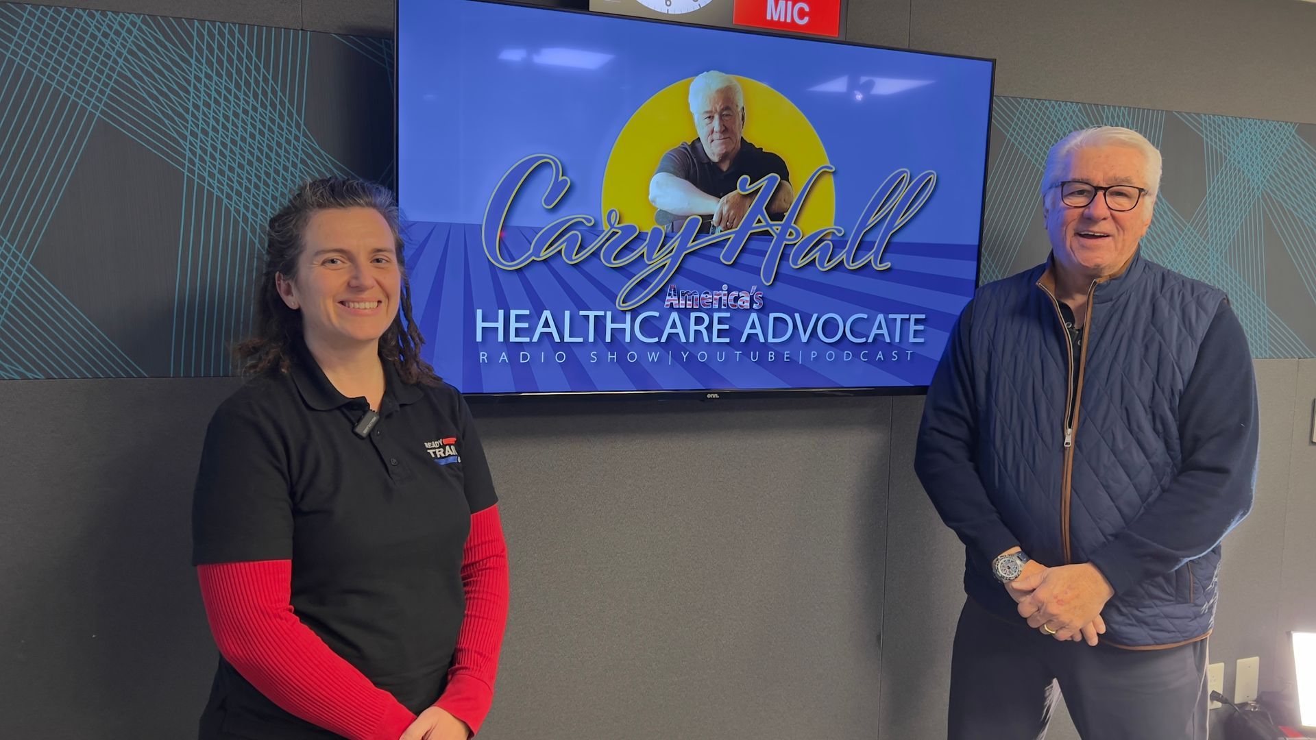 Two people standing beside a wall-mounted “Health Care Advocate” sign in an indoor office setting.