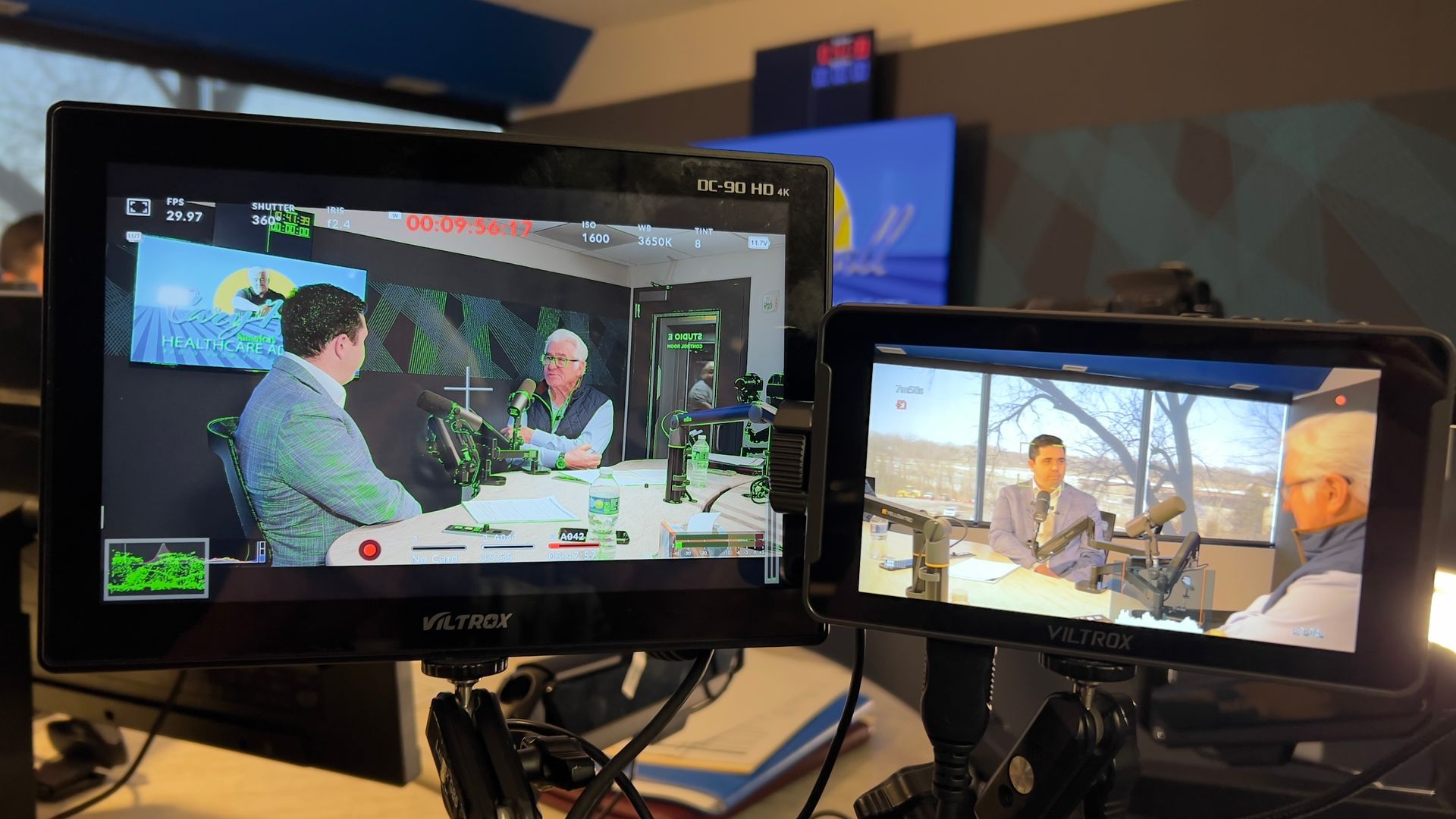 Two monitors showing a video call in a studio, with two people seated at a table on the left screen and outdoors on the right