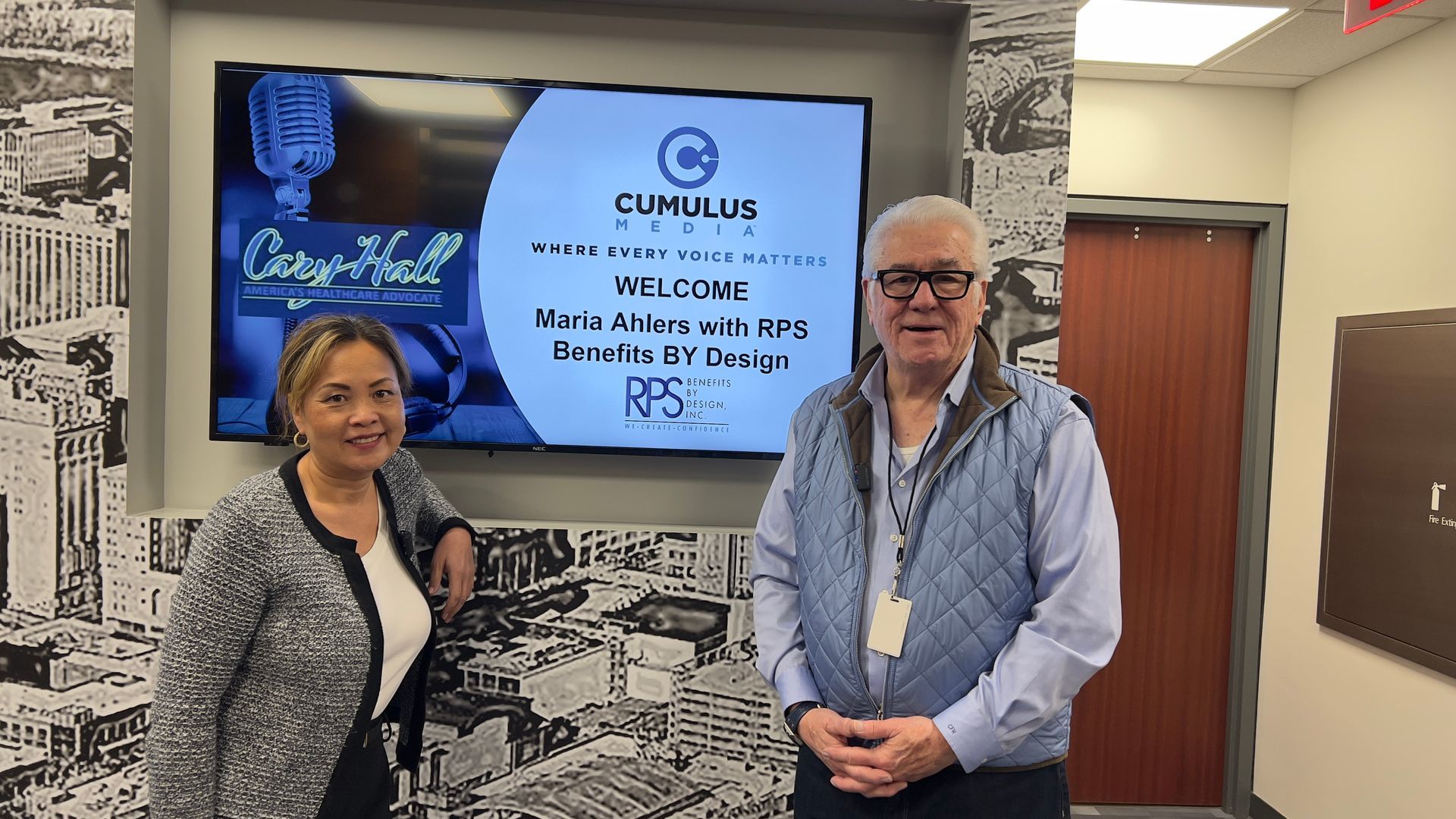 Two people standing by a wall-mounted screen in an office hallway, with a Cumulus logo and display behind them.