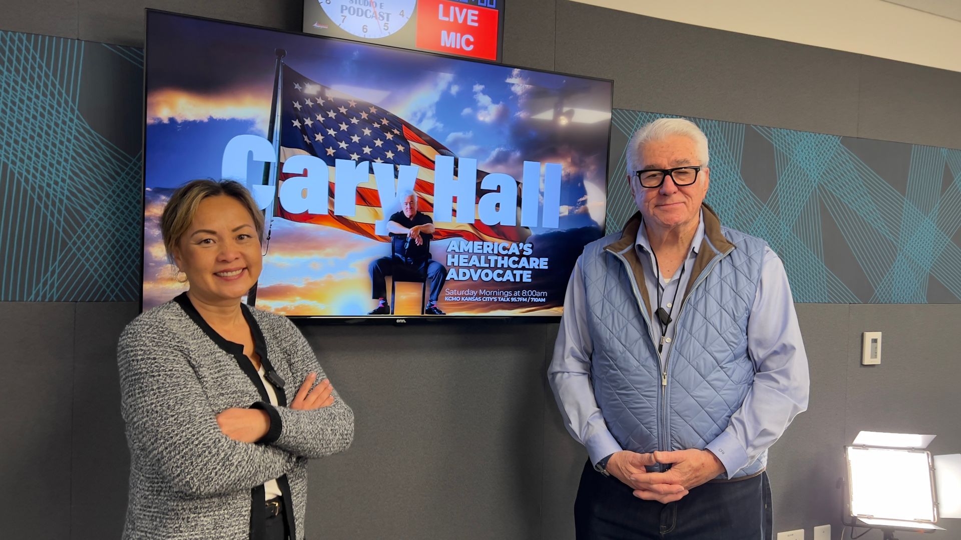 Two people standing in front of a TV screen displaying the Gary Hall Live logo.