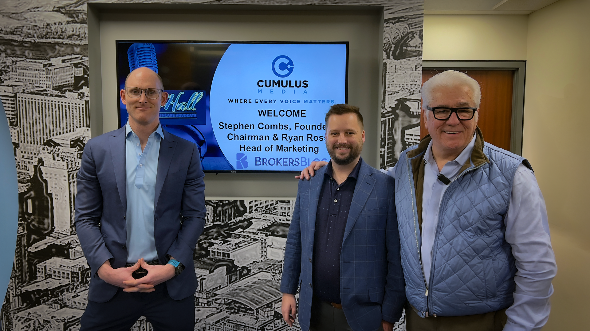 Three men standing in an office lobby beside a screen with the Curl/Science logo and meeting information.