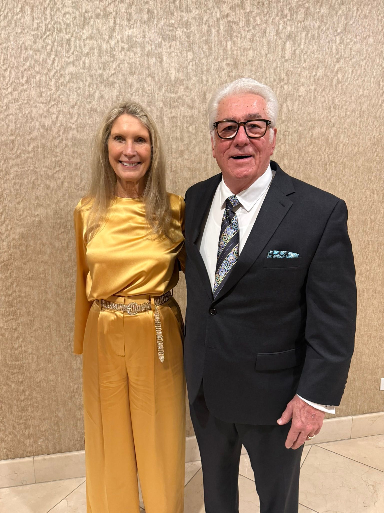 Two people posing indoors, wearing formal attire, in front of a textured wall.