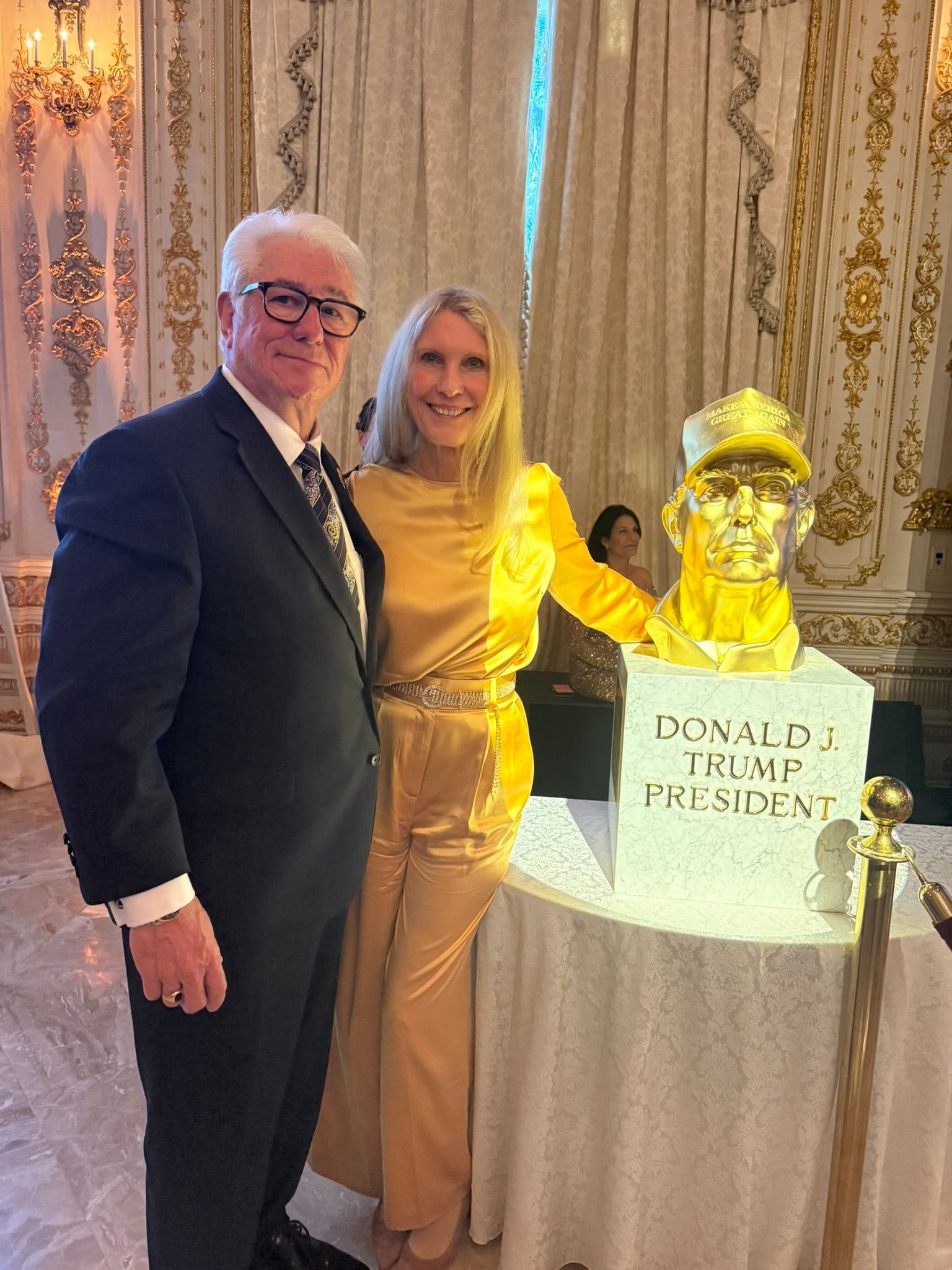 Two people posing beside a gold-lit Donald Trump cake at an indoor event