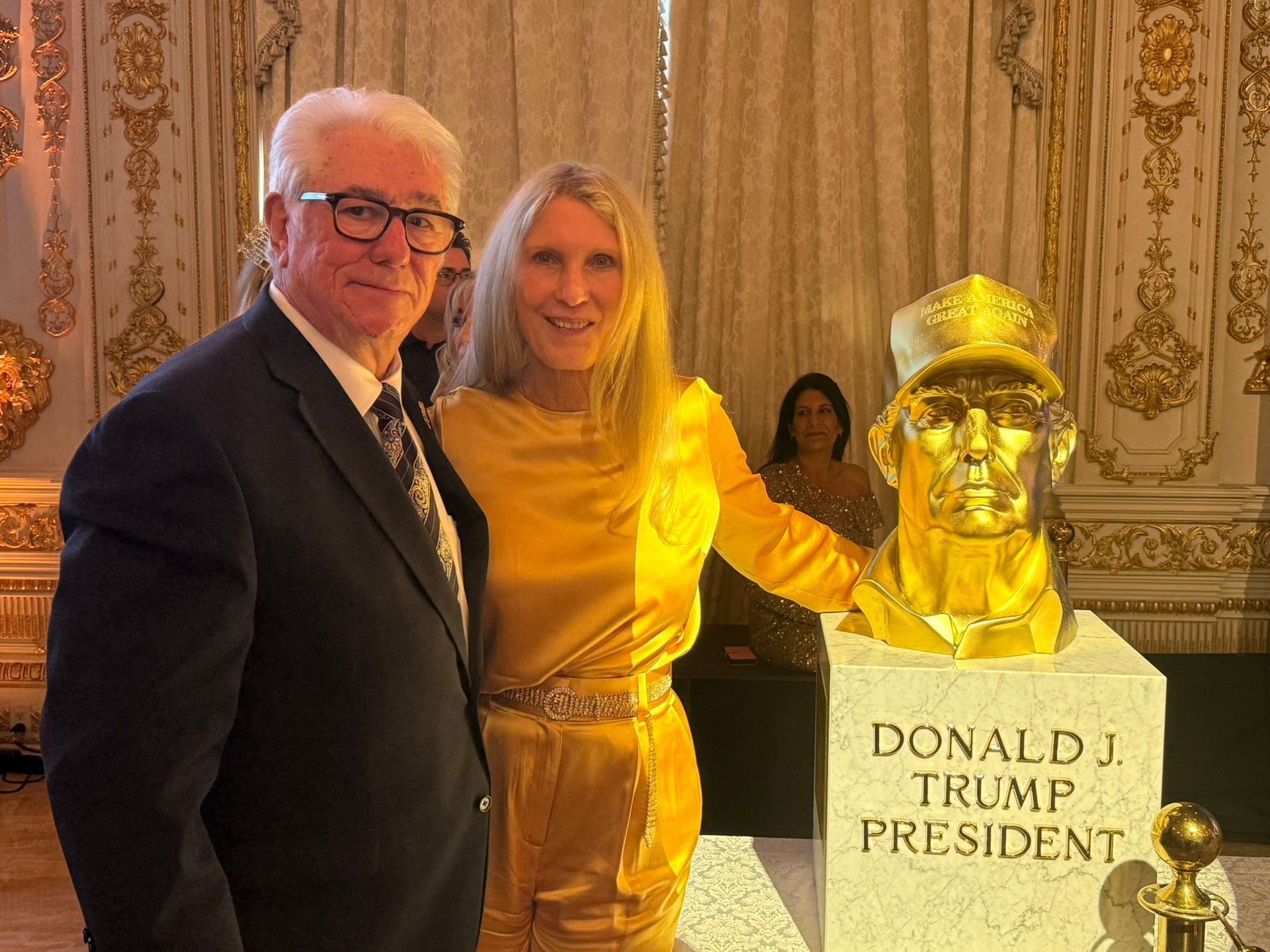 Two people posing beside a gilded bust labeled “Donald J. Trump” in an ornate room.