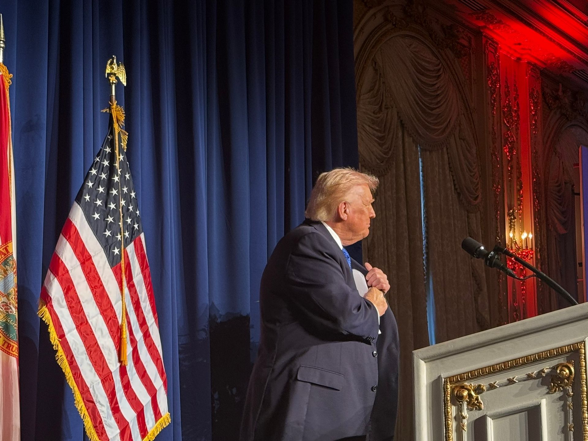 Speaker at podium beside U.S. flag, adjusting his tie on a stage with blue curtains and red lighting