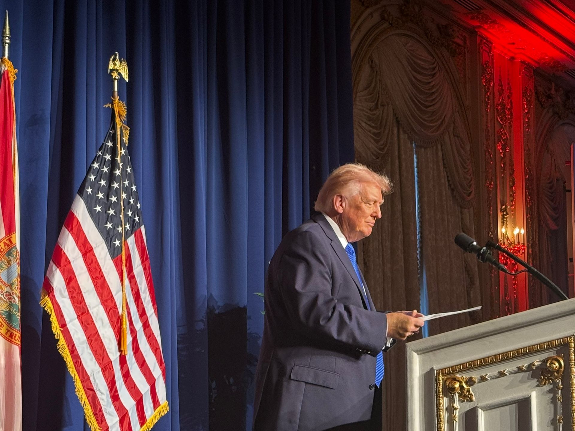 Donald Trump speaking at a podium beside a U.S. flag on a stage with blue curtains and red lighting
