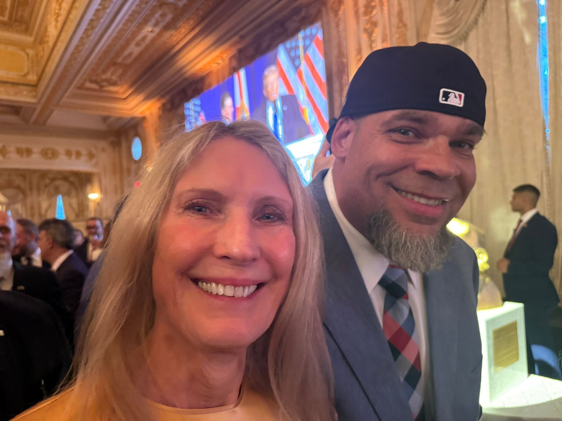 Two people smiling at an ornate indoor event, one in a suit and cap, with a large screen in the background