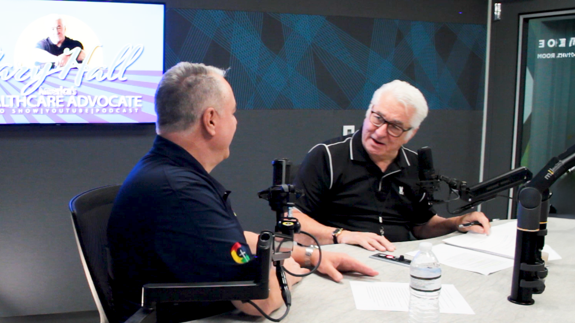 Two men in a podcast studio, speaking across a table with microphones and headphones.