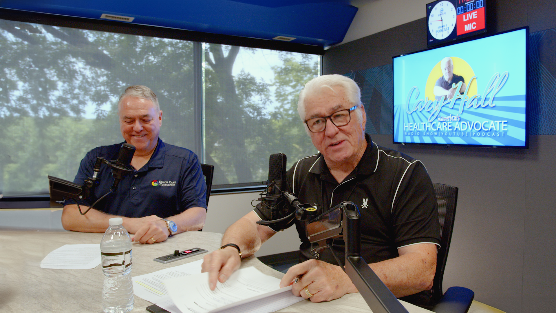 Two men sit at a conference table with a microphone and papers, speaking in a meeting room.
