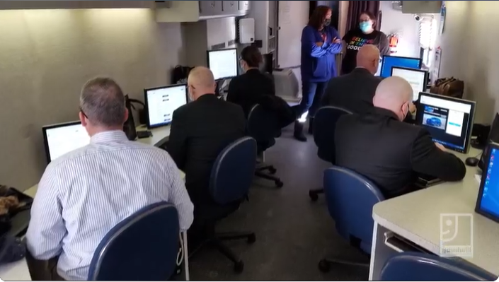 People seated at computers in a small classroom, watching a presentation at the front.
