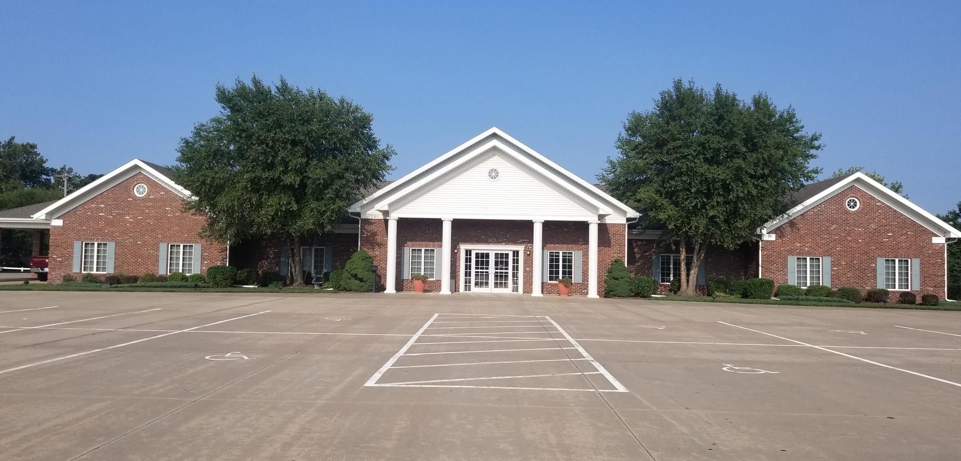 Brick building with white trim, trees, and a parking lot. Clear blue sky.