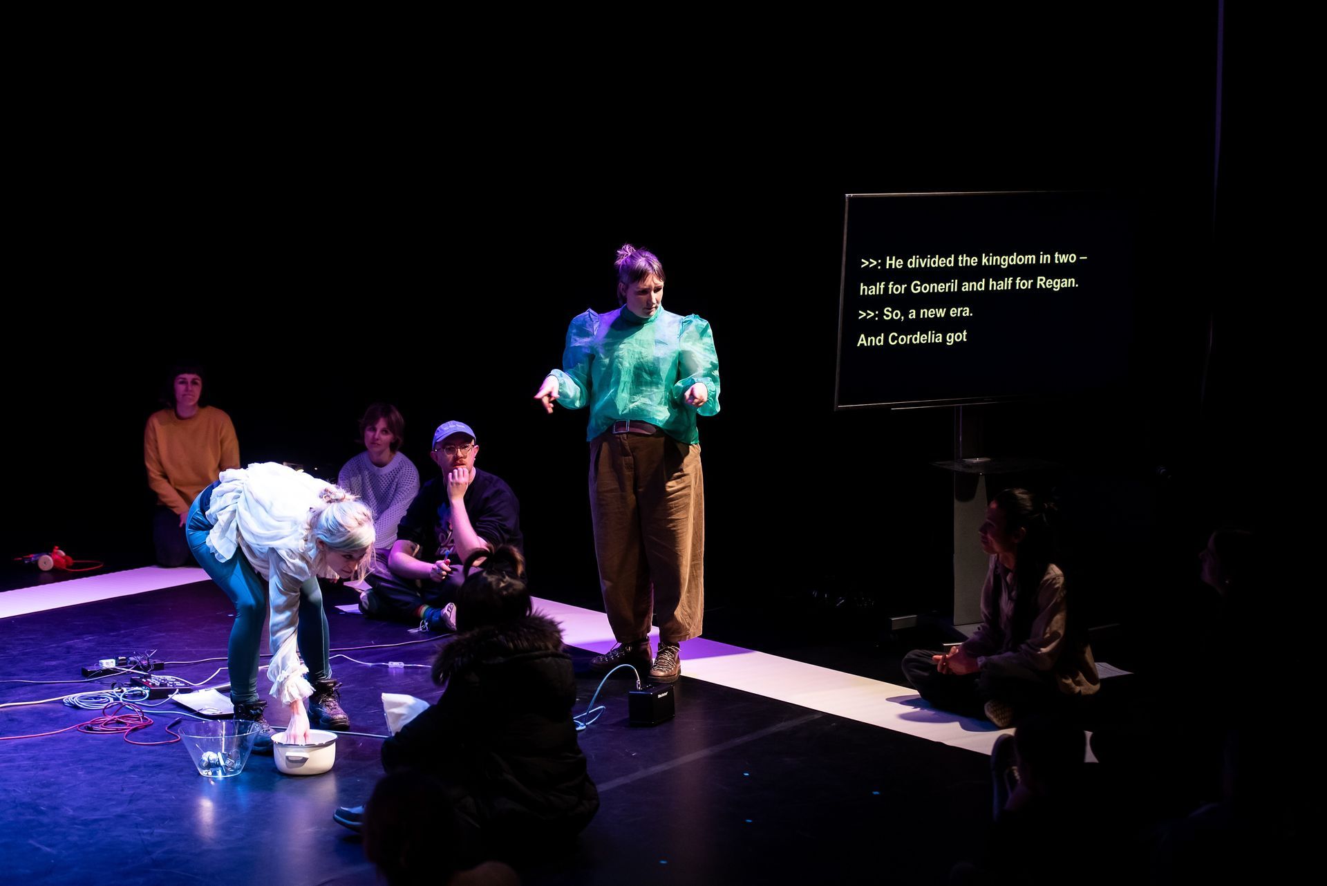 Performer on stage, points to something in foreground with both hands and a quizzical look. They are surrounded by seated audience members on the stage. To one side, is a television screen with yellow captions.
