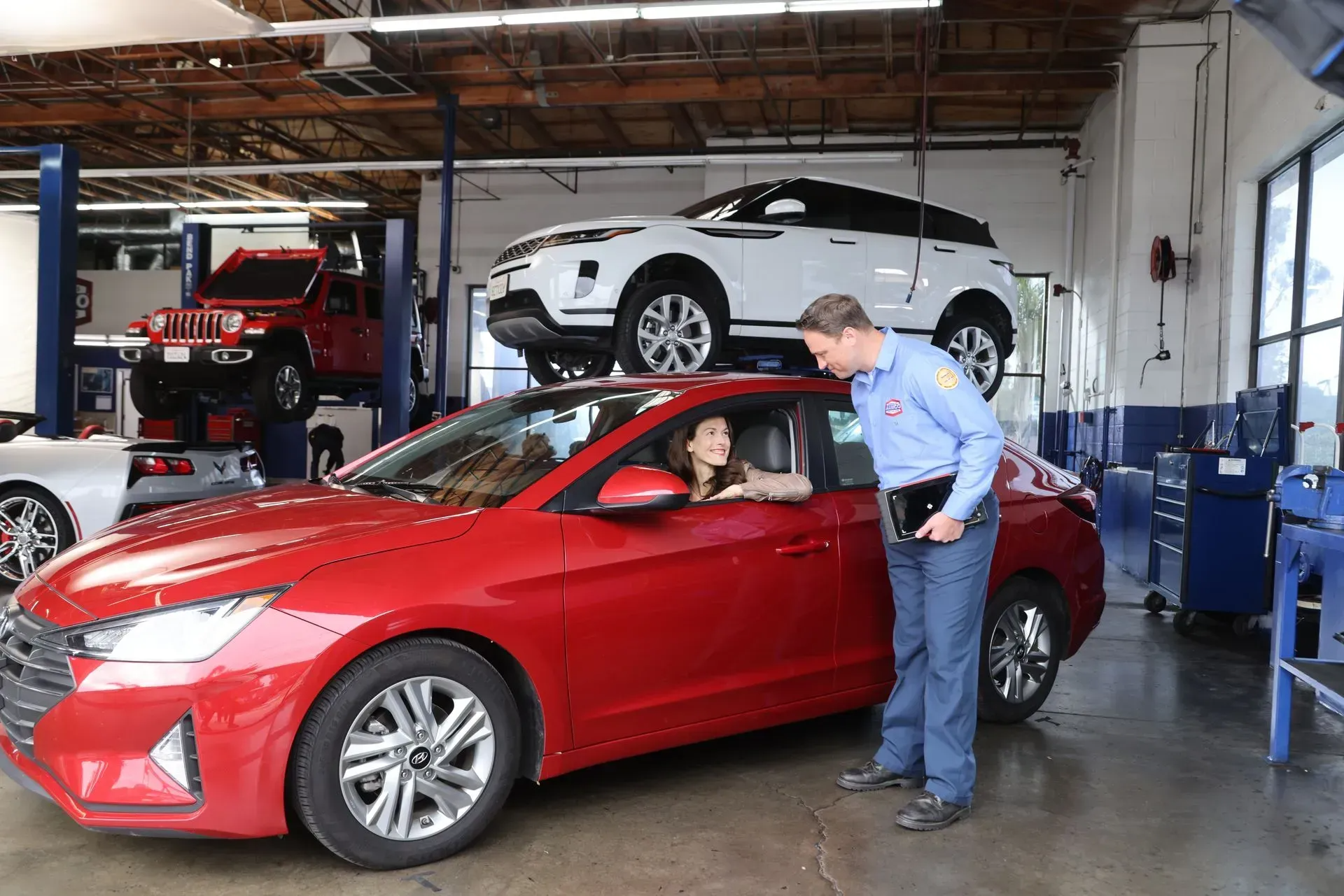 An AAMCO mechanic talks to a customer in a red car at a repair shop, other cars on lifts.