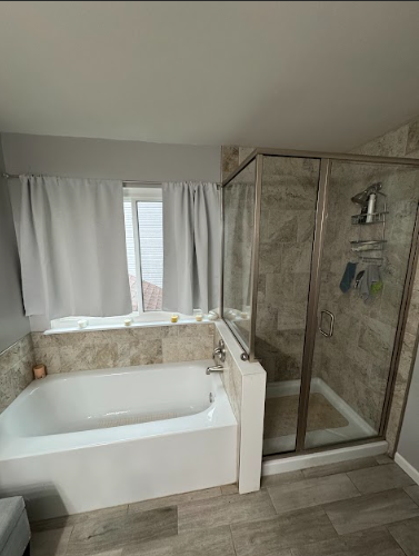 A modern bathroom with a white bathtub, a glass shower, and a window with gray curtains.