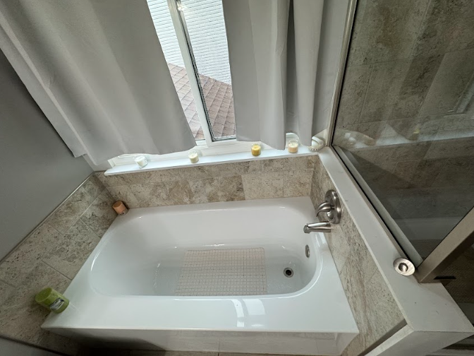 White bathtub with tiled surround next to a window with white curtains. Shower door is to the right.