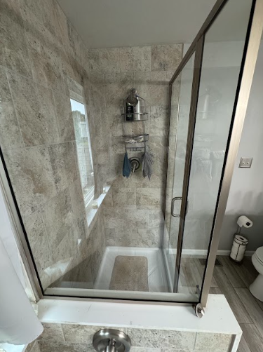 A well-lit, tiled shower stall with a glass door. There are shelves for toiletries, towels, and a rug inside.
