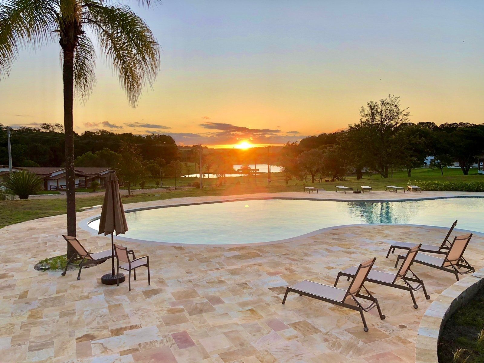 Um pôr do sol sobre uma grande piscina com cadeiras e guarda-sóis.
