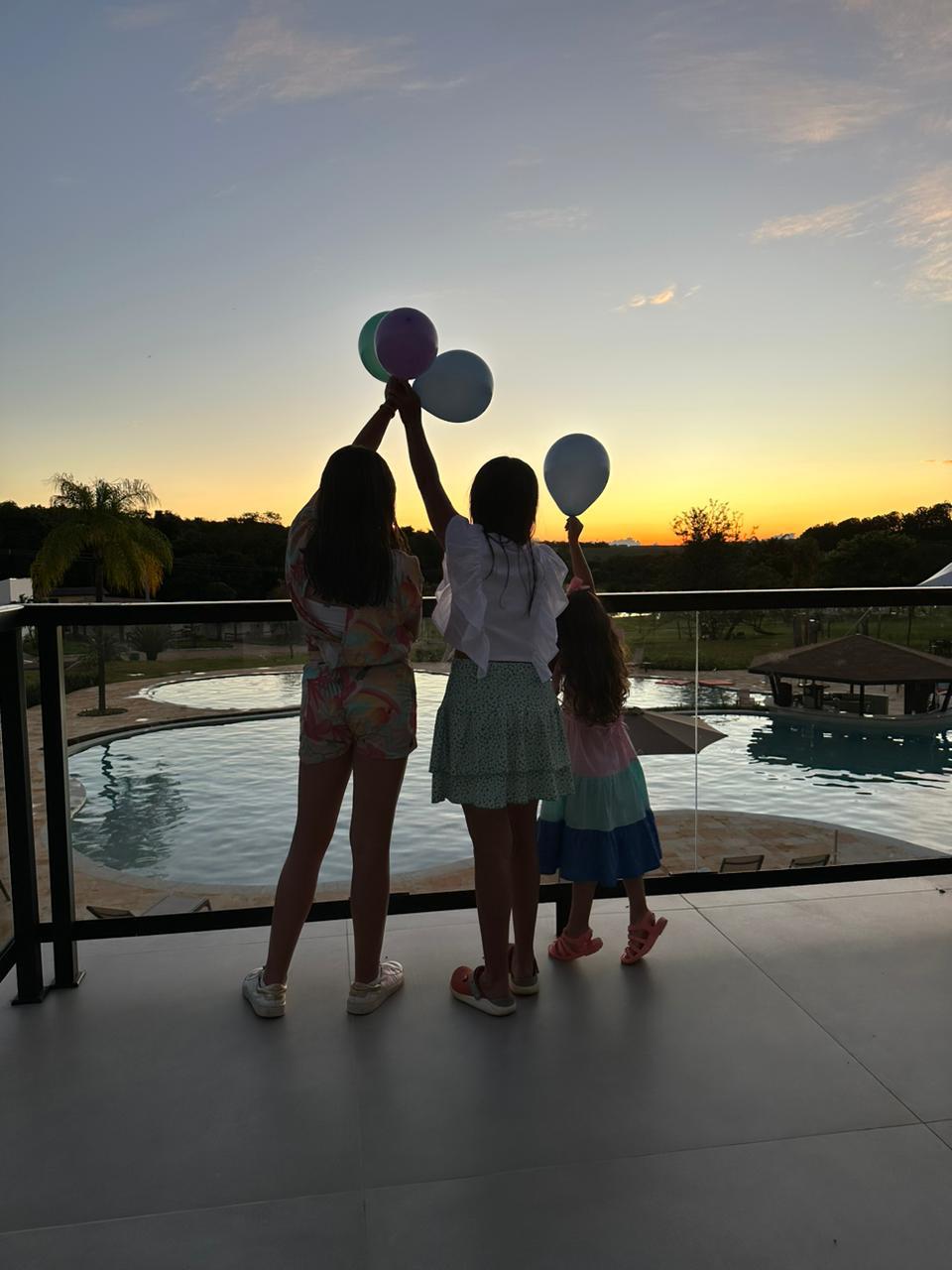 Três meninas estão em uma sacada segurando balões ao pôr do sol.