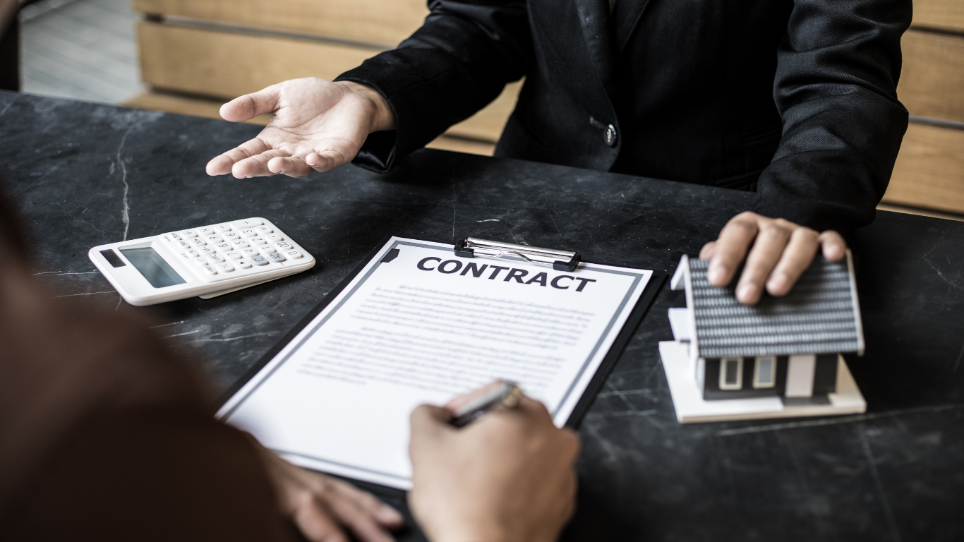 Person signing a contract, with a miniature house, calculator, and another person at a table.