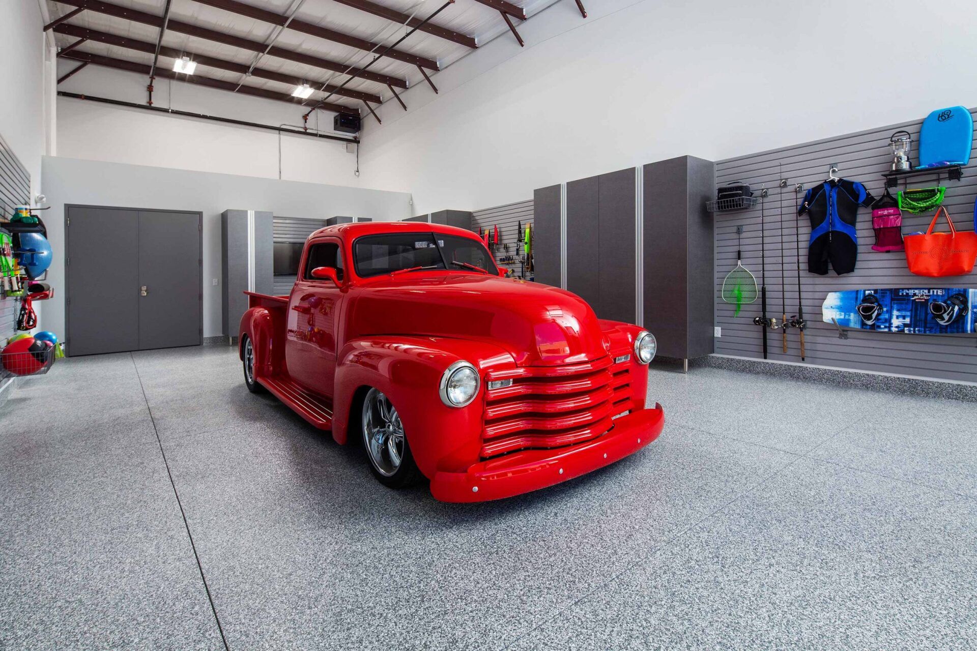Red classic pickup truck in a modern garage with gray walls and epoxy flooring, sporting gear visible.