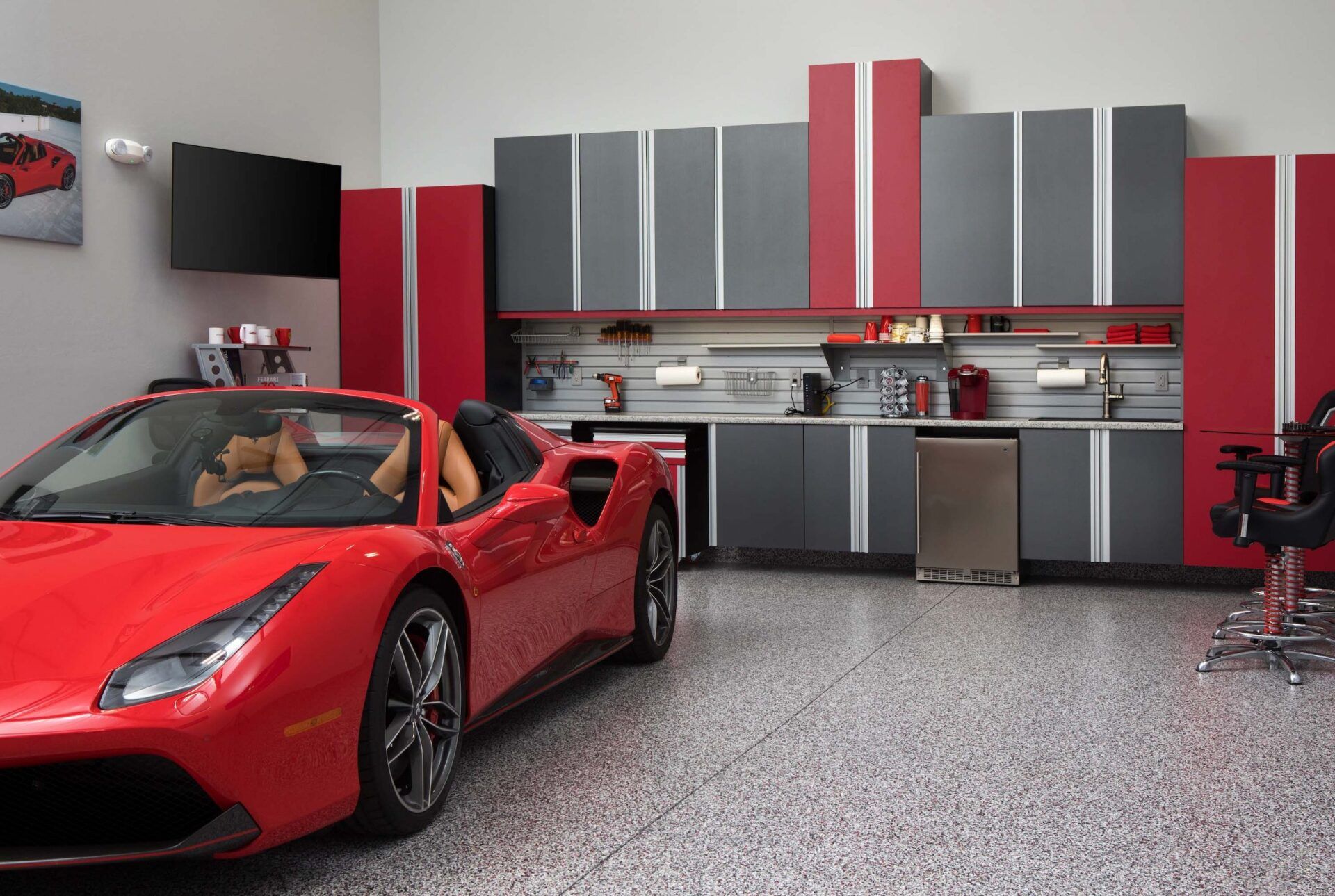 Red sports car parked in a garage with grey and red cabinets, a TV, and a shiny, speckled floor.