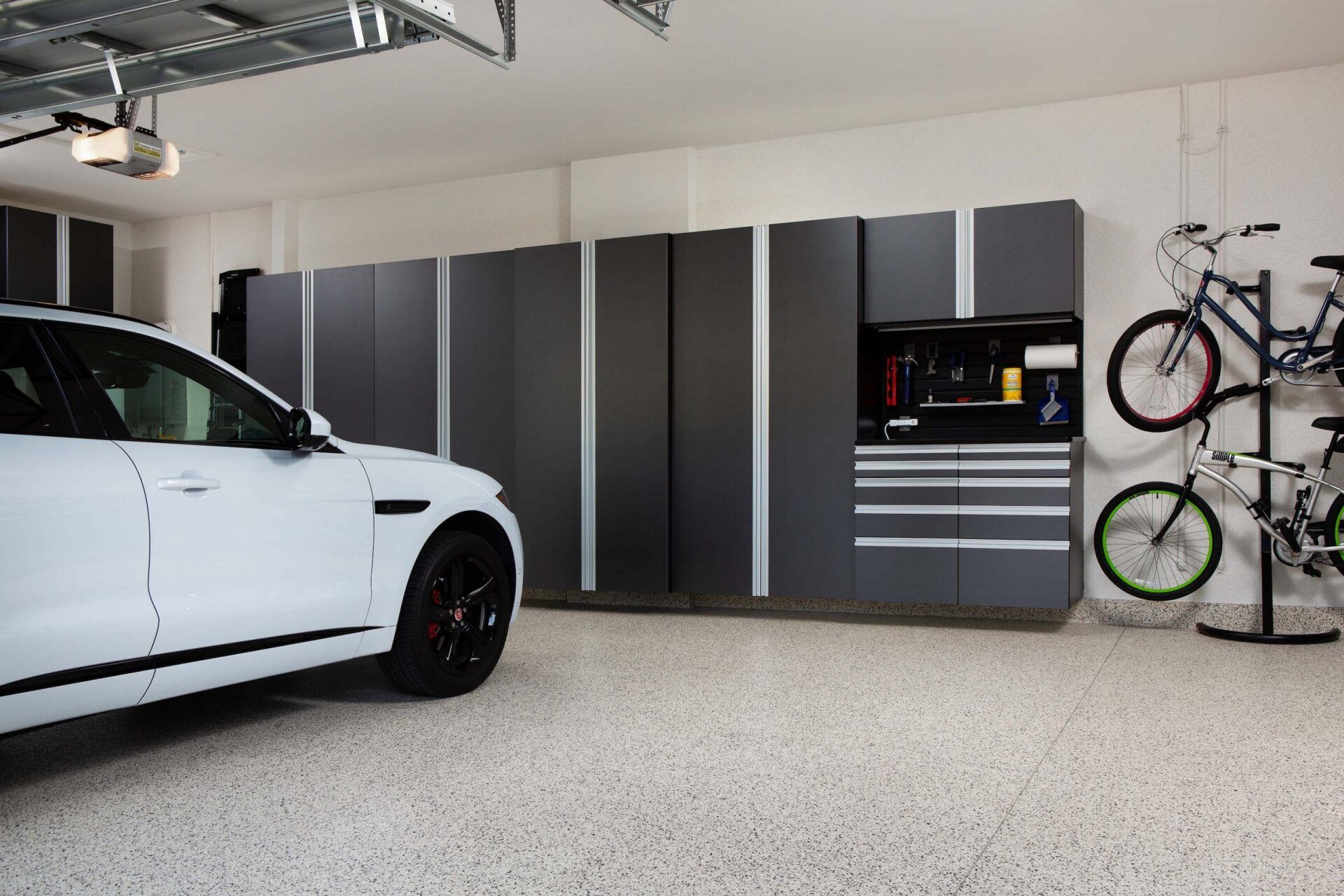 White car parked in a garage with dark gray storage cabinets and two bikes.