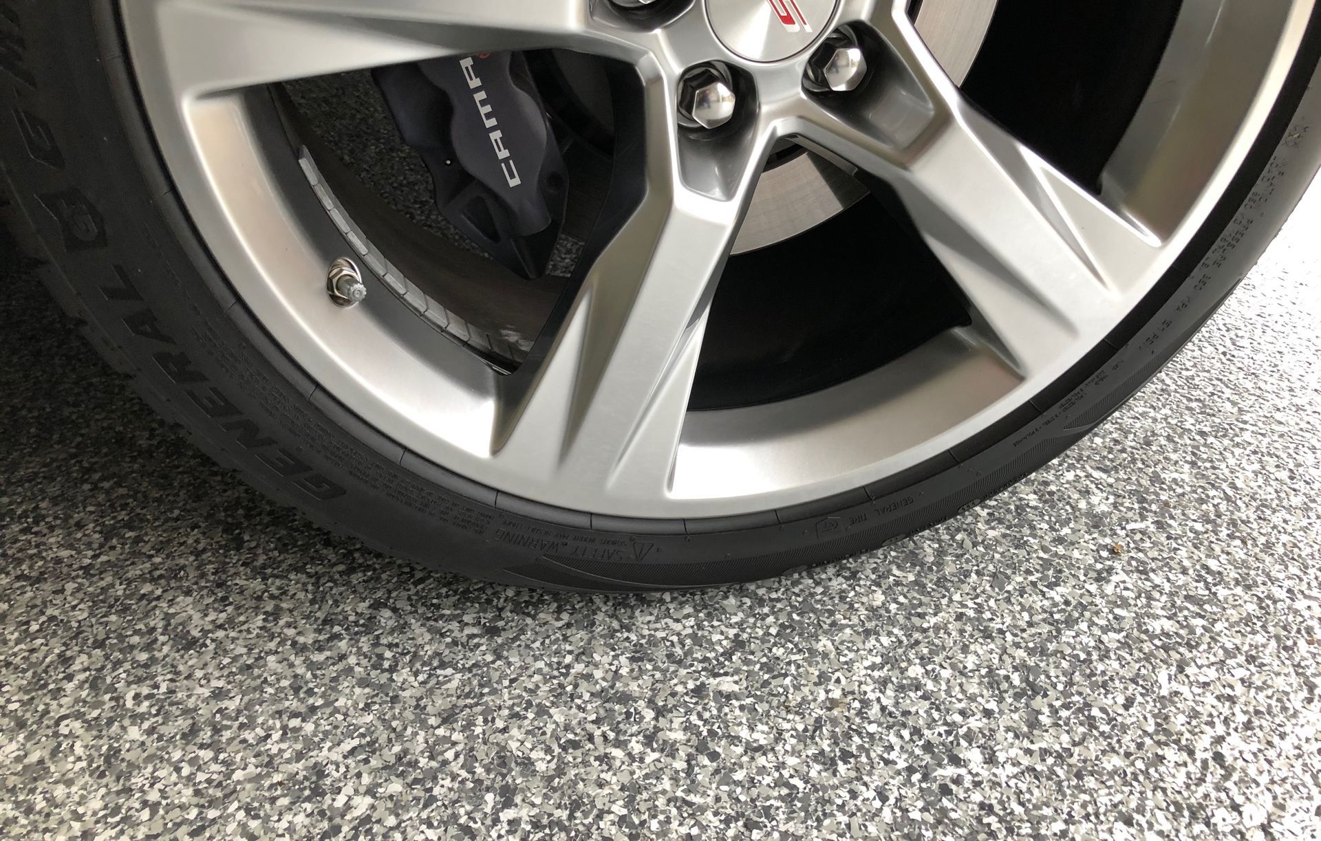 Silver car wheel with black tire on speckled gray floor.