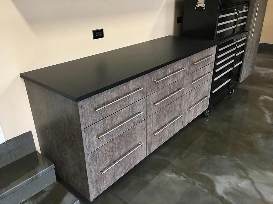 Garage cabinets with dark countertop and gray faux wood grain drawers; adjacent to black tool cabinet.