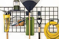 Black metal grid wall organizer holding gardening tools. Yellow hose and tools, green rake, and olive green lantern.