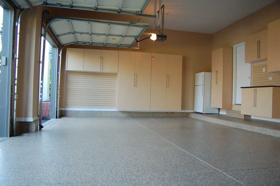 An empty garage with a speckled floor and tan cabinets. The garage door is open.