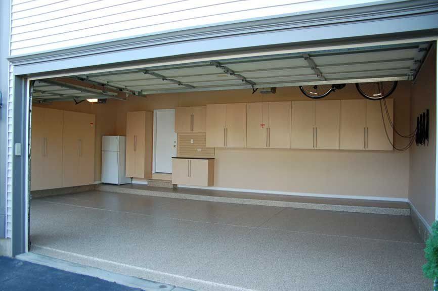 An open garage with tan walls, cabinets, and a gray epoxy floor. A bicycle hangs from the ceiling.